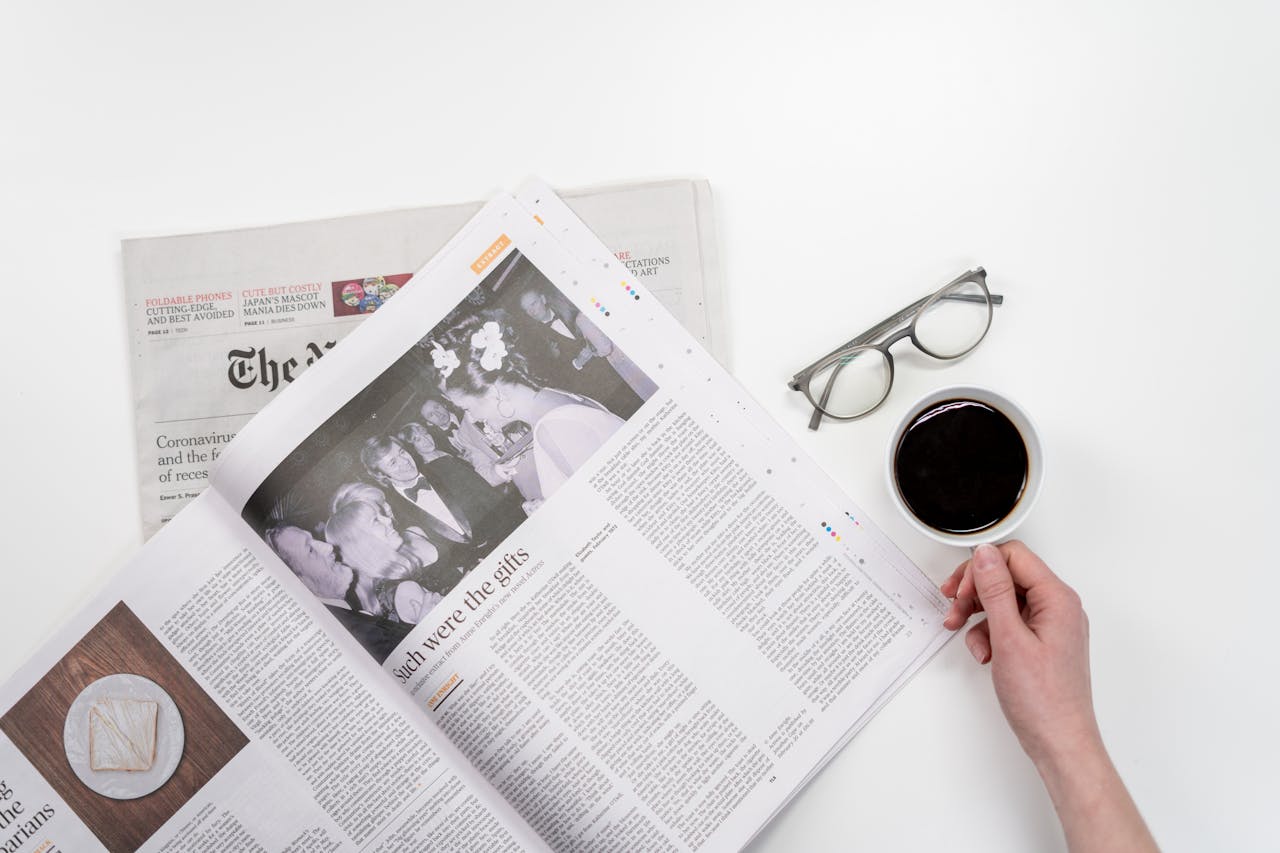 Overhead view of newspaper reading with coffee and glasses on a white table.